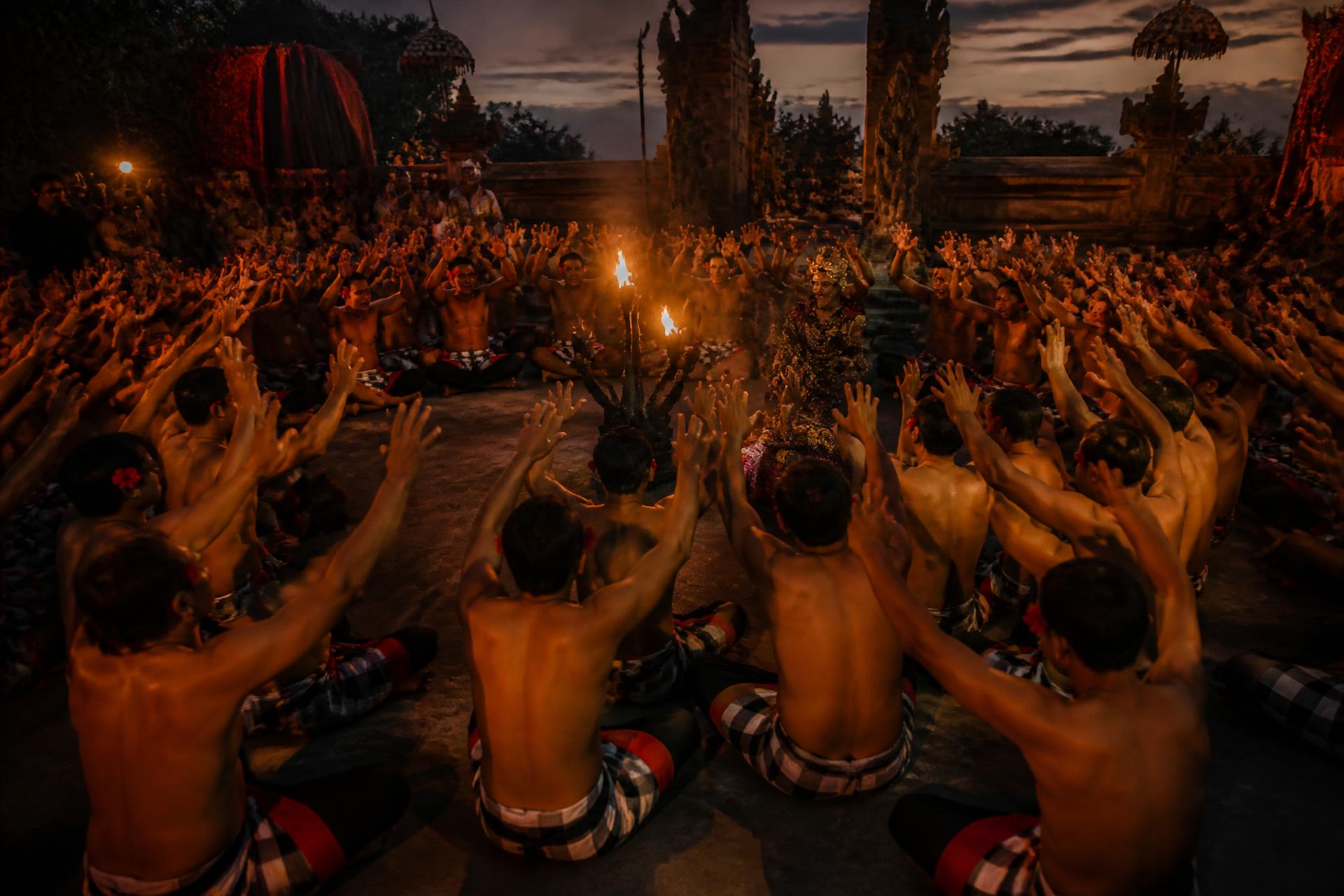 Performers seated in a wide circle at dusk with arms raised around a central flame, in a Balinese temple courtyard